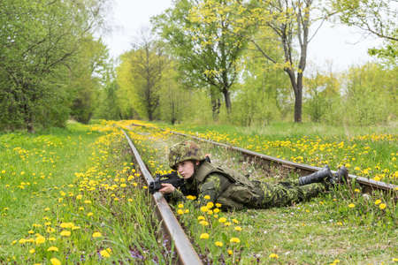 Portrait of armed woman with camouflage. Young female soldier observe with firearm on rail. Child soldier with gun in war, green grass and yellow dandelions background.  Military, army people conceptの写真素材