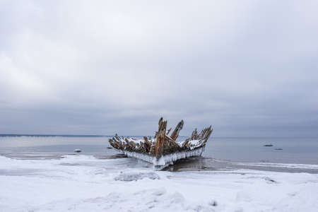 Old broken boat wreck and rocky beach in wintertime. Frozen sea, evening light and icy weather on shore like fairy tale country. Winter on coast. Blue sky, white snow, ice covers the land.の写真素材