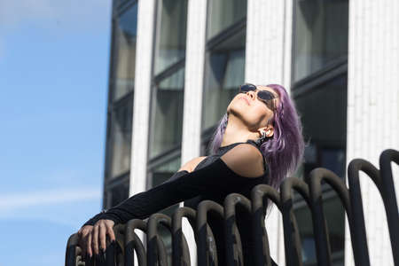 Caucasian white female model standing on the balcony of the building and enjoys weather. Beautiful woman with long purple hair, in a gray dress resting in the city, modern fashion concept.の写真素材