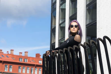 Caucasian white female model standing on the balcony of the building and enjoys weather. Beautiful woman with long purple hair, in a gray dress resting in the city, modern fashion concept.の写真素材