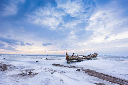 Old broken boat wreck and rocky beach in wintertime. Frozen sea, evening light and icy weather on shore like fairy tale country. Winter on coast. Blue sky, white snow, ice covers the land.の写真素材