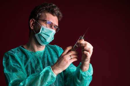 Doctor in medical mask make protective injection. Nurse in glasses prepare syringe. Medic preparing vaccination against for a pandemic and flu season. Dentist in disposable medical clothing.の写真素材
