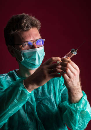 Doctor in medical mask make protective injection. Nurse in glasses prepare syringe. Medic preparing vaccination against for a pandemic and flu season. Dentist in disposable medical clothing.の写真素材