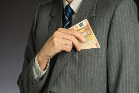 Businessman in suit putting banknotes in his jacket breast pocket. Business man is holding cash, stack of fifty euros money. Person pays in euro bills, isolated gray background.の写真素材