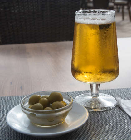 Olives in a small bowl and a glass of ice cold beer on a table の写真素材