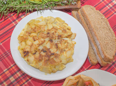 Plate with homemade tortilla de patata, rosemary herbs and slices of bread on red tablecloth の写真素材