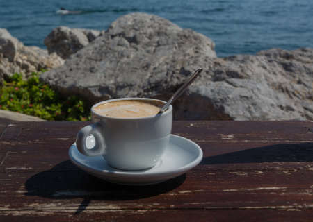 Cappuccino in white cup and spoon on weathered wooden table and ocean in the background の写真素材