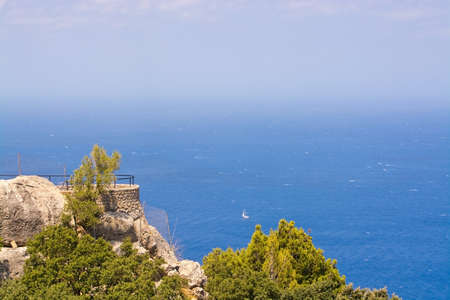 Mediterranean ocean scenery with stone terrace, sailboat and white seafoam on a sunny windy day in Mallorca, Balearic islands, Spain in July.の写真素材