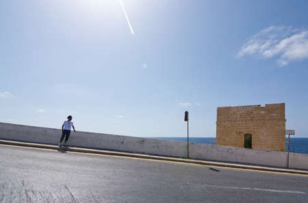 BLUE GROTTO, MALTA - SEPTEMBER 15, 2015: Maltese bus driver rests near the downhill bus stop with stone building, ocean horizon and airplane contrail in backlight, of popular tourist attraction Blue Grotto on a sunny day in September 15, 2015 in Malta.のeditorial素材
