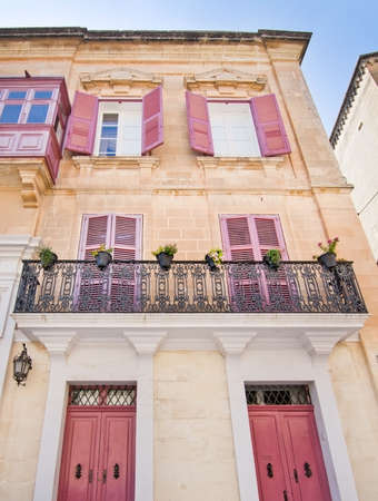 MDINA, MALTA - SEPTEMBER 15, 2015: Building with red window shutters and black wrought iron balcony inside old city walls on a sunny day in September 14, 2015 in Mdina, Malta.のeditorial素材
