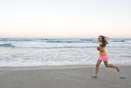 CANYAMEL, MALLORCA, BALEARIC ISLANDS, SPAIN - JULY 18, 2016: Young smiling happy brown haired girl runs barefoot speed blur on the sandy beach with ocean waves, pink sunset horizon and full moon on a summer evening on July 18, 2016 in Canyamel, Mallorca, のeditorial素材