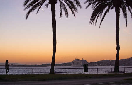 PALMA DE MALLORCA, SPAIN - JANUARY 4, 2018: Paseo Maritimo with silhouette palm trees, woman walking and ferry boat at dusk on January 4, 2018 in Palma de Mallorca, Spain.のeditorial素材