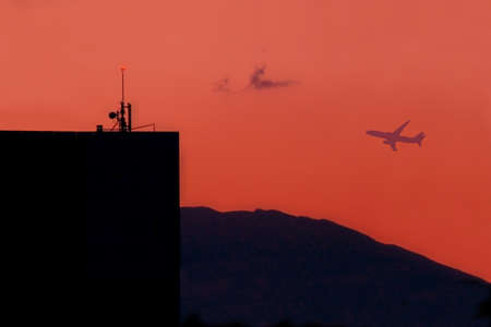Aircraft silhouette takes off near building with antenna and safety lights on red evening sky,...