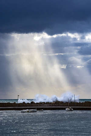 Vertical Mediterranean sea view with pier against dark skies with dramatic light on splashing waves on a winter day in Mallorca, Spain.の写真素材