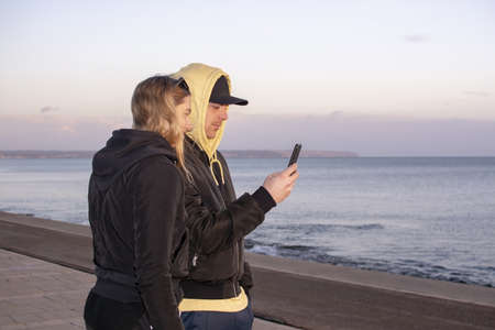 Handsome young natural and casual looking couple with hood jackets black and yellow watch sunset and...