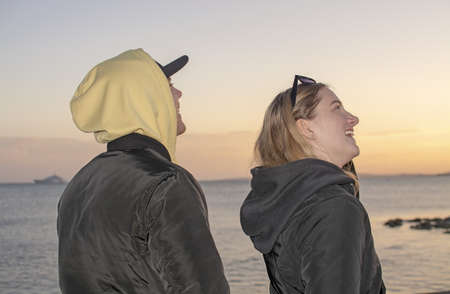 Closeup shot of handsome young natural and casual looking couple with black and yellow hood jackets laughing and watch up towards yellow sunset sky on a beach.の写真素材