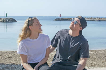 Young natural smiling and casual sporty couple sit on stone wall in sunshine by the ocean in...