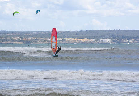 CAN PASTILLA, MALLORCA, SPAIN - APRIL 6, 2019: Windsurfer with red sail plays in the green waves on a windy and sunny day on April 6, 2019 in Can Pastilla, Mallorca, Spain.のeditorial素材