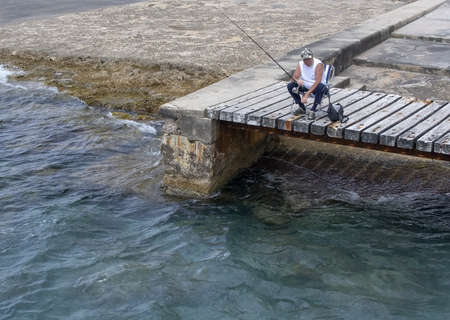 SANTA PONSA, MALLORCA, SPAIN - MAY 29, 2019: Relaxed fisherman sitting on a wooden bridge by the water on May 29, 2019 in Santa Ponsa, Mallorca, Spain.のeditorial素材