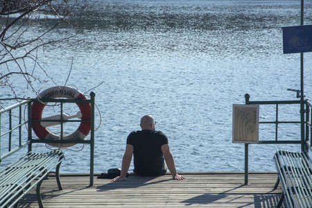 STOCKHOLM, SWEDEN - APRIL 20, 2019: , Man sitting on the dock by a lake in stark spring sunshine on...
