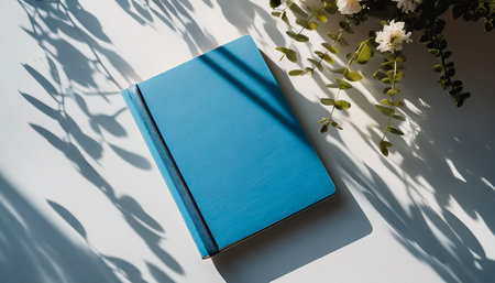 closed notebook with blue blank cover mock up on white table with natural floral sun light shadows aesthetic minimalist company business branding templateの素材