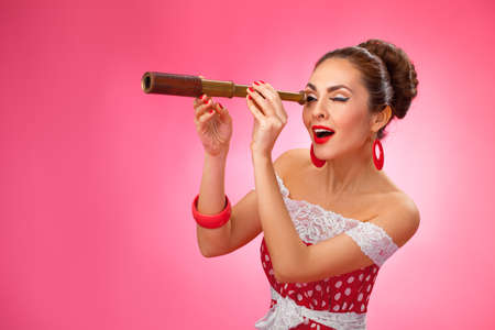 Woman is looking through a telescope and making a surprised expression. She is standing against a pink background.の写真素材