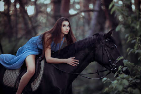 Young beautiful brunette girl in blue dress ride on the black horse in forestの写真素材