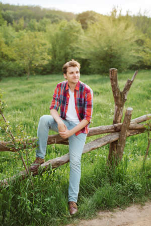 Young smile man. Gorgeous guy in casual clothes, sit on a fence, rural look , outdoorsの写真素材