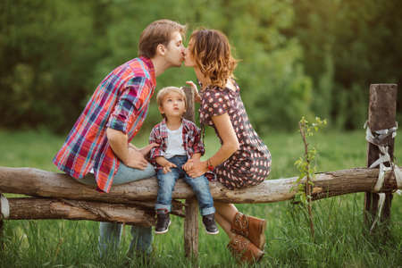 Happy Family on the nature. Mother father kissing and in casual clothes, sit on a fence, rural look , outdoorsの写真素材