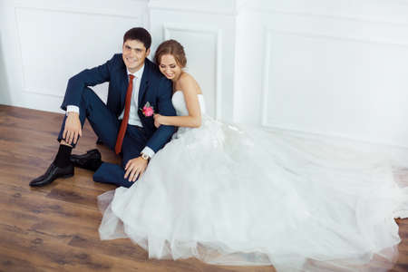 Wedding couple in love. Beautiful bride in white dress with brides bouquet and handsome groom in blue suite sitting on the floor and embracing each other indoors in decorated studio room, white bright interior with big window. Young fashion models, real cの写真素材