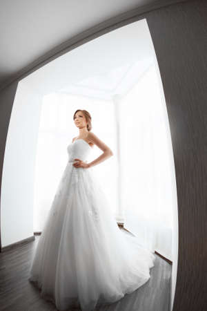 Beautiful young bride in white wedding dress indoors.  Luxuty model standing in full length against big window at home. Girl showing fashion look of wedding dress in bright room. High key, made on fisheye lens photo.の写真素材