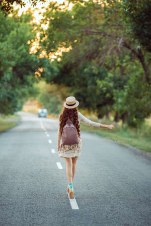 Hitchhiking tourism concept. Travel hitchhiker woman with hat and backpack walking on road during holiday travelの写真素材