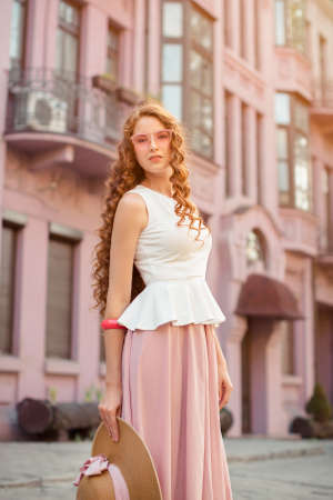 Beautiful urban girl. Fashion pretty woman wearing a retro elegant hat, sunglasses, pink skirt and handbag walking in city over grey buildings.の写真素材