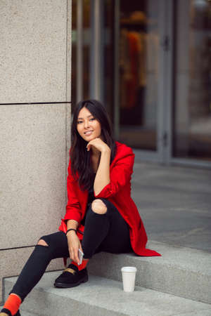 Happy asian businesswoman smiling in the city. Happy lovely and beautiful mixed race Asian Caucasian young woman in casual red suite outdoor sitting on steps of building with coffee cup. Next generationの写真素材