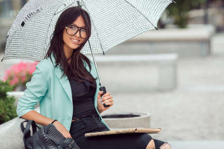 Asian girl city portrait. Woman smiling and sitting on bench outside with umbrella in casual business suite. Beautiful young mixed race Asian Caucasian woman.の写真素材