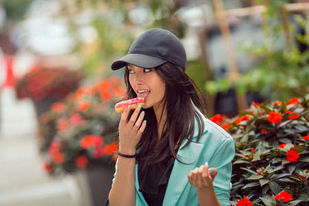 Asian beautiful woman eating a donut sitting near bed of flowers on street, fast food concept outdoors. Busy businessman have no time.の写真素材
