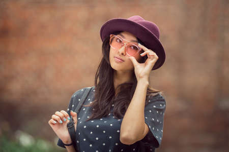Asian student girl portrait. Beautiful woman holding glasses against brick wall bokeh background. Mixed race student girl in university college campus park serious looking at camera.の写真素材