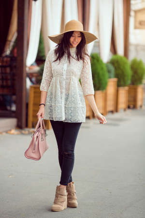 Elegant Asian woman in white dress with hat on the street. Girl standing in full length outside with...