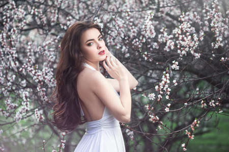 Sensual young woman standing in white dress at blossoming tree in the garden. Beauty of woman and nature consept. Portrait of beautiful model with curly brown hair posing and looking at camera.の写真素材
