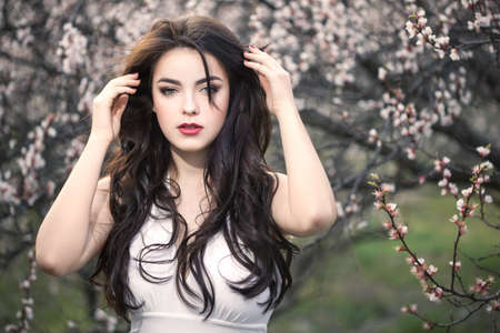 Sensual young woman standing in white dress at blossoming tree in the garden. Beauty of woman and nature consept. Portrait of beautiful model with curly brown hair posing and looking at camera.の写真素材