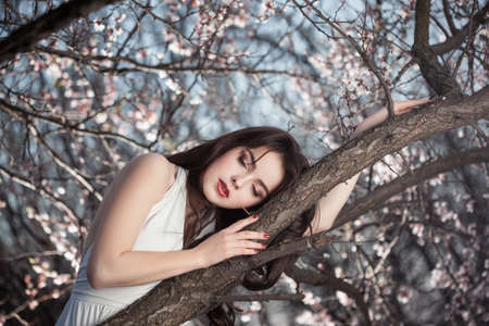 Sleeping beauty. Young gorgeous woman with in white dress perfect make-up lying on spring tree branch with flowers blossom. People save the nature concept. Perfect model with closed eyes.の写真素材
