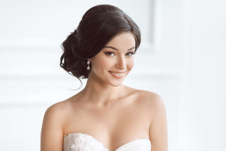 Beauty portrait of bride wearing wedding dress with feathers with luxury delight make-up and hairstyle, studio indoor photo. Young attractive multi-racial Asian Caucasian model. Smiling beautiful young woman like a bride in white room at home looking at cの写真素材