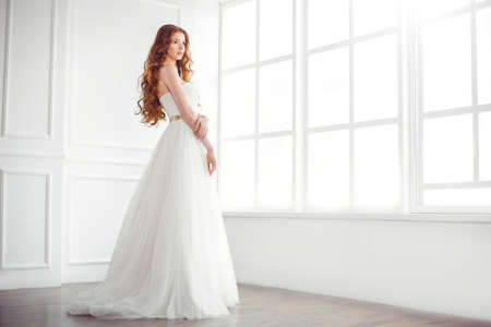 Wedding. Beautiful bride indoors at big window in full lenght looking away. Side viewの写真素材