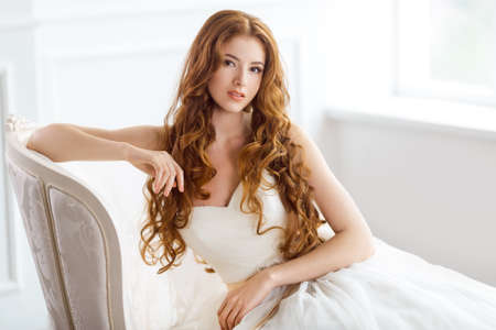 Bride in beautiful dress sitting on sofa indoors in white studio interior like at home. Trendy wedding style shot in full length. Young attractive caucasian red hair model like a bride against big window tender resting.の写真素材