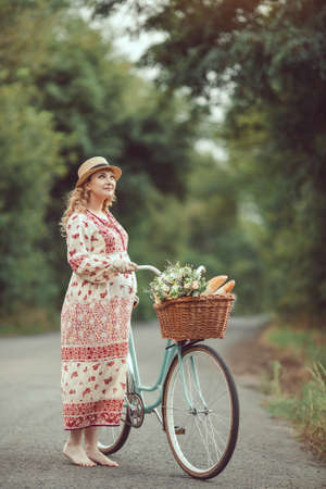 Pregnant girl retro French style with bicycle on a forest road. Beautiful pregnancy concept. Blonde happy woman with curly hair on nature background.の写真素材