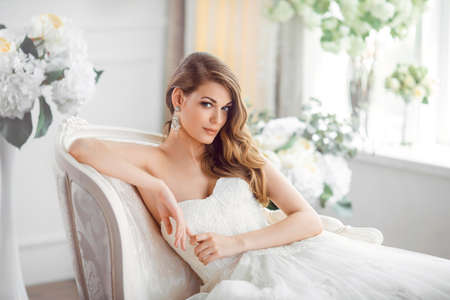 Wedding. Bride in beautiful dress sitting on sofa indoors in white studio interior like at home. Trendy wedding style shot. Young attractive caucasian brunette model like a bride against big window tender posing.の写真素材