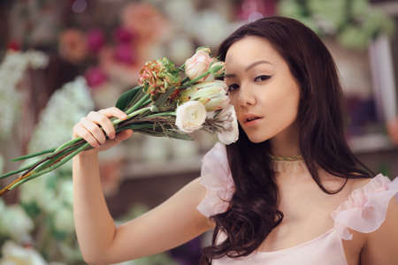 Beautiful girl in tender pink dress with bouquet flowers peonies in hands standing against floral bokeh background in flower shop. Joyful asian female holding bouquet near face. Playful fashion model looking at cameraの写真素材