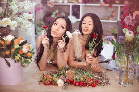 Beautiful asian florist girls making bouquet of flowers on table for sale against floral background in flower shop indoors. Two attractive asian females florists working in store. 2 playful fashion models in tender dresses posing playing with flowers.の写真素材