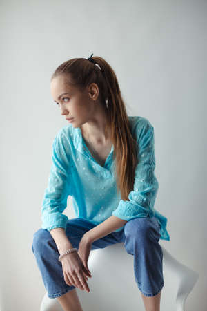 Confident stylish urban young woman looking away while sitting on chair, on gray background. Closeup fashion studio portrait of hipster young tender woman in shirt and jeans. Caucasian teen woman model posing indoorsの写真素材