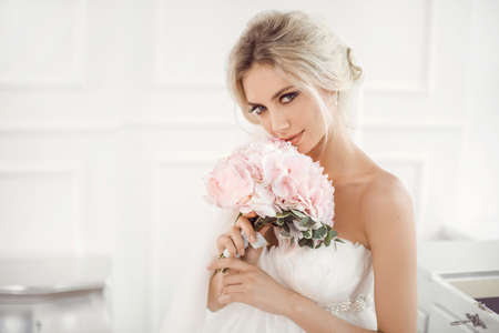 Classical young gourgeous bride. Studio interior fashion shot of fashion model in wedding dress with bouquet of flowers sitting in white room. Blonde woman portait with profeshional make-up and hairstyle.の写真素材